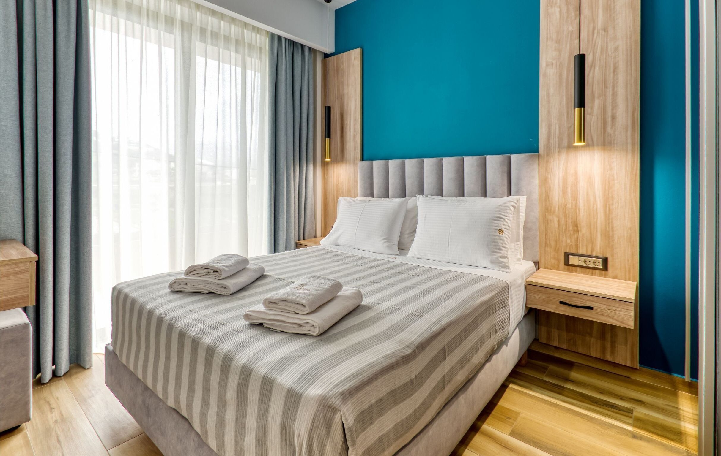 Ein modernes Hotelzimmer mit einem großen Bett, grauen Vorhängen, einem Holz-Kopfteil und einer blauen Akzentwand.