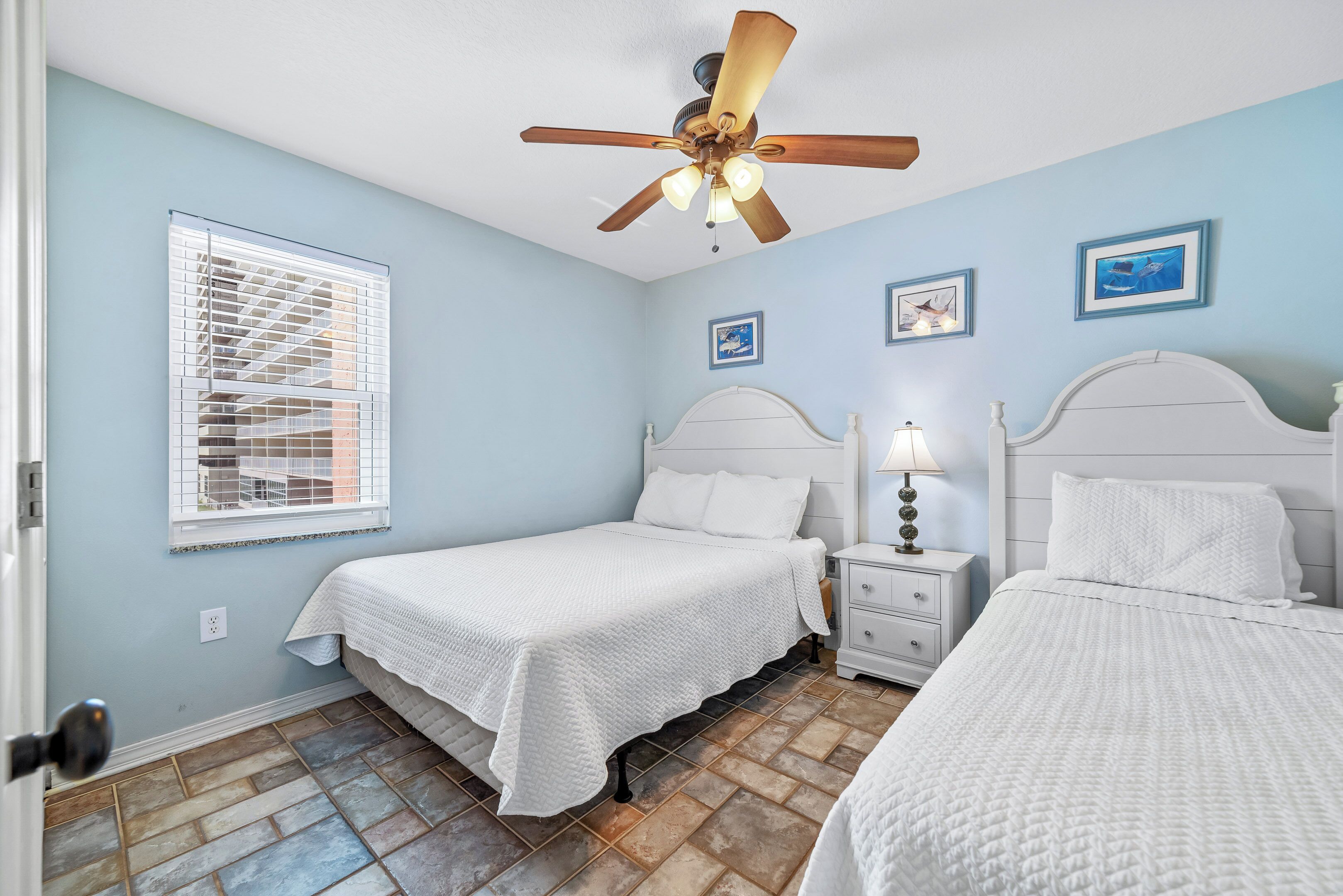 A bedroom with two beds, a ceiling fan, a nightstand with a lamp, and a window with blinds.