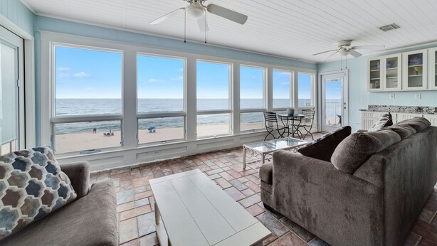 Ein geräumiges Zimmer mit großem Fenster zum Strand, Deckenventilator und einer Sitzecke mit Sofa und Tisch.