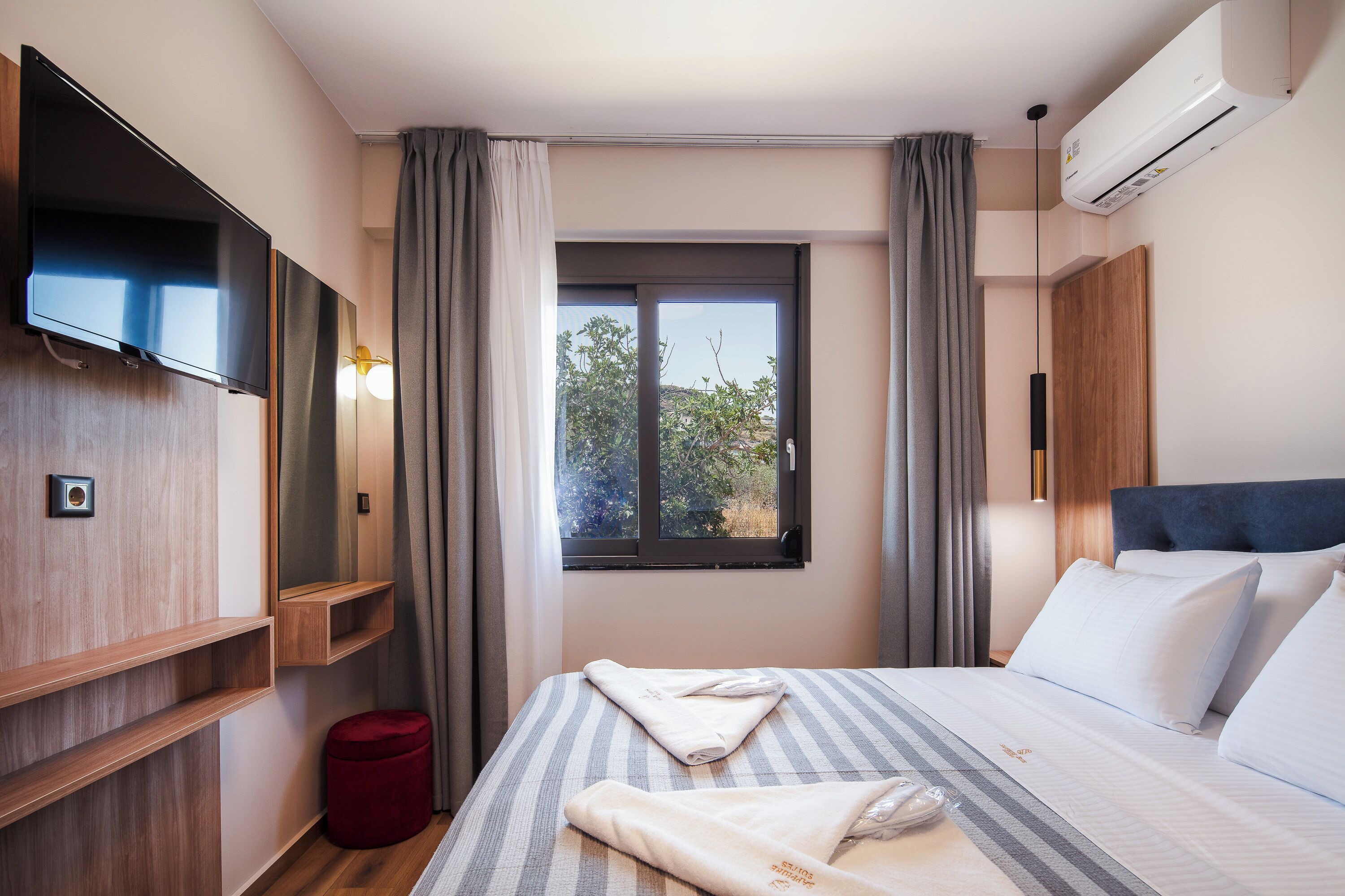 A hotel room with a bed, a TV, a window with curtains, an air conditioner, and a small red stool.