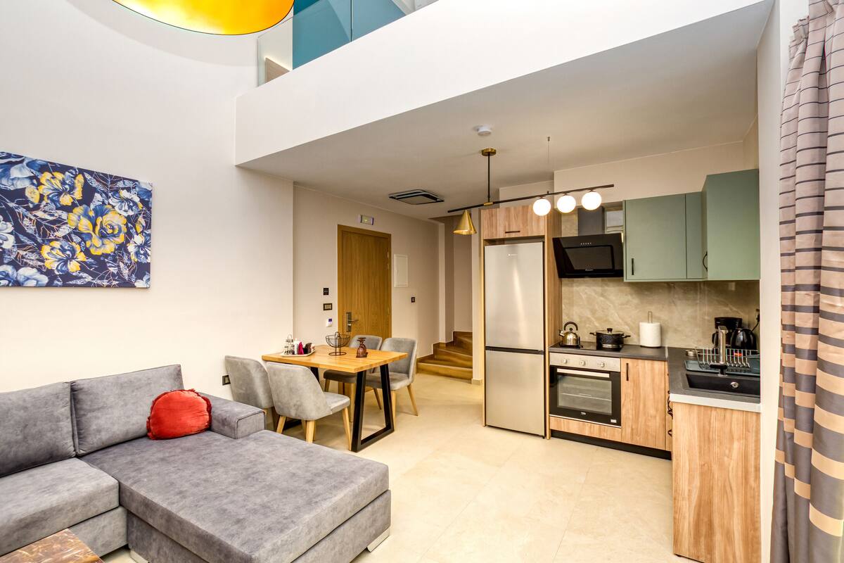 A modern apartment with a grey sofa, a dining table, a kitchen with a refrigerator, stove, and microwave.
