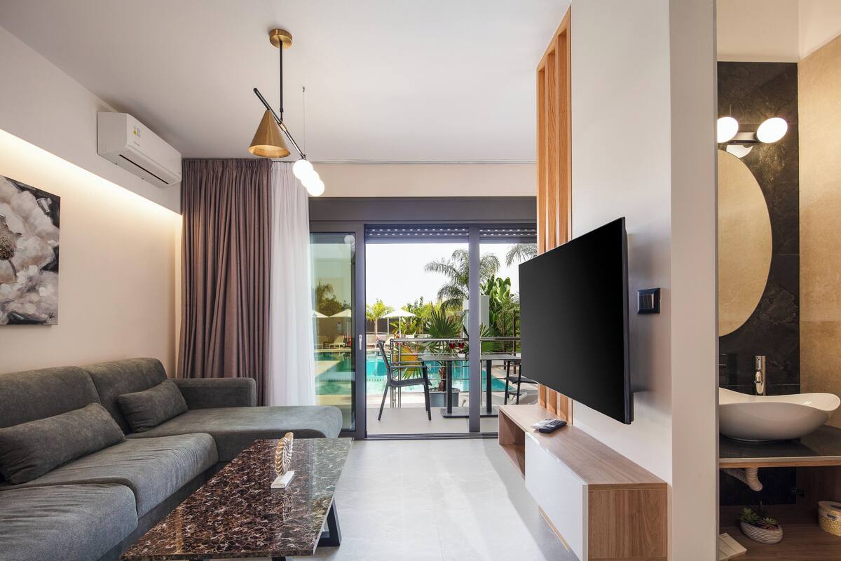 A modern living room with a grey sofa, a marble coffee table, a flat-screen TV, and a large window overlooking a pool area.