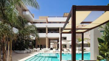 Area piscina con lettini e ombrelloni, circondata da palme e un edificio a più piani.