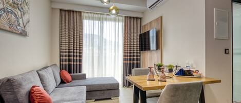 A modern living room with a grey sofa, a wooden dining table, and a grey chair. There is a TV mounted on the wall and an air conditioning unit above it.