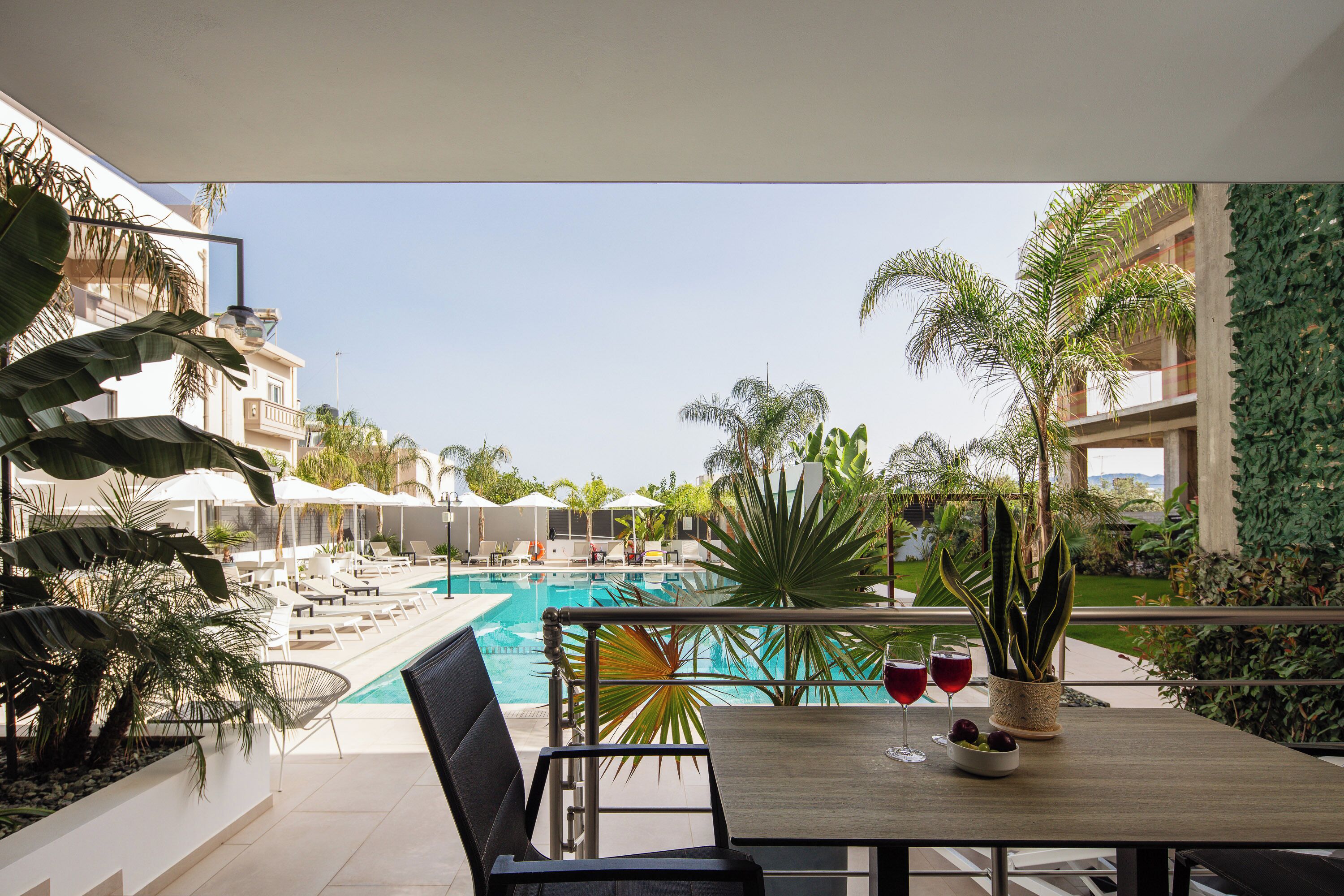 Area piscina con sedie a sdraio, un tavolo da pranzo con due bicchieri di vino e vista su una piscina e vegetazione.