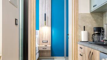 Un pasillo estrecho que conduce a un dormitorio con una pared azul decorativa, una mesita de noche con lámpara y una pequeña cocina con cafetera y escurreplatos.