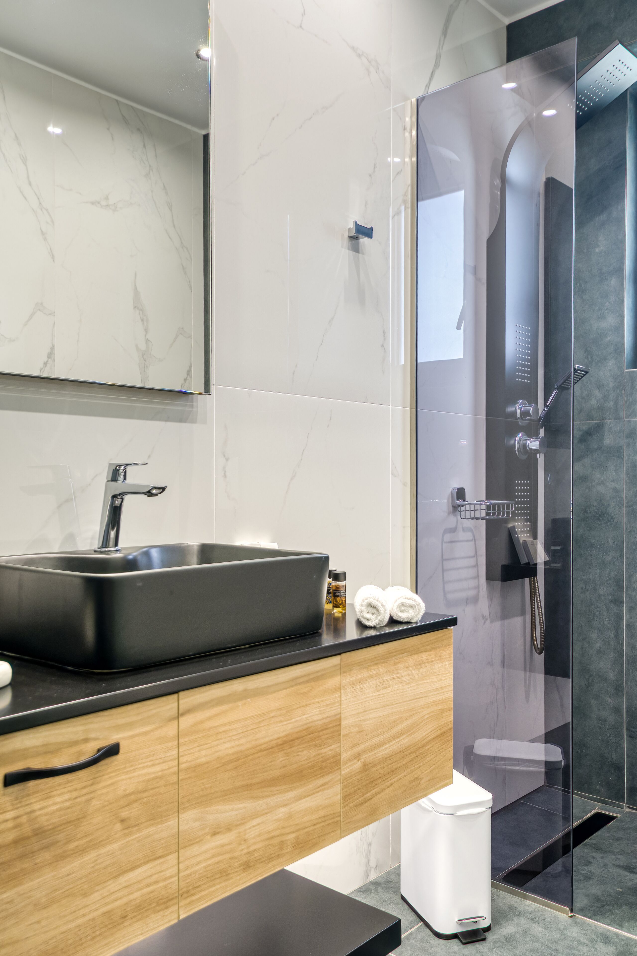 A modern bathroom with a large sink, wooden cabinets, a glass shower enclosure, and a marble-tiled wall.