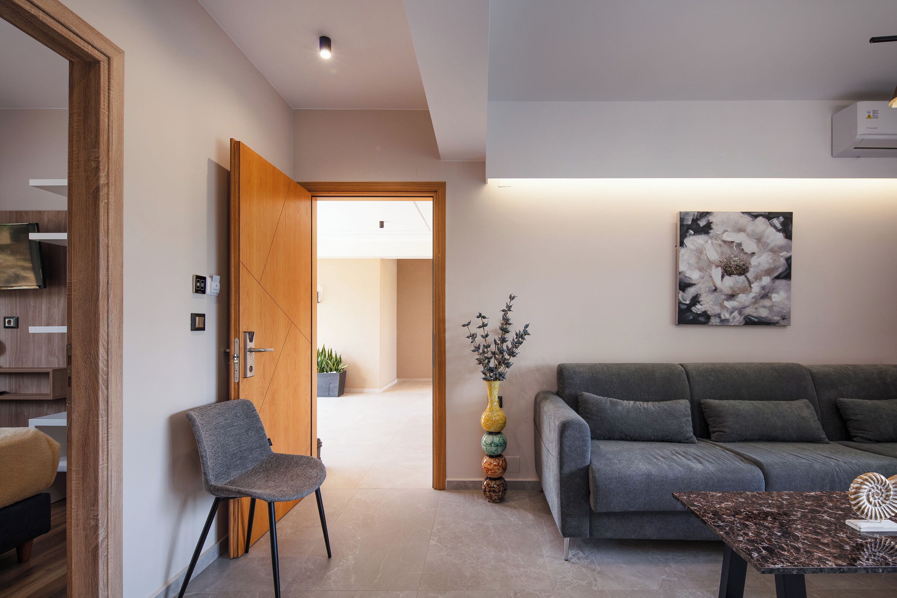 Uma sala de estar moderna com um sofá cinzento, uma mesa pequena e uma cadeira. Uma porta de madeira dá acesso a outro cômodo, e há uma obra de arte na parede acima do sofá.