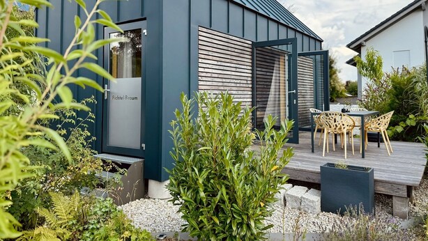 A modern building with a dark facade, a wooden slatted door, and a patio area with outdoor furniture.