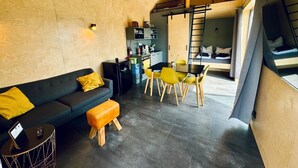 A compact living space with a sofa, ottoman, dining table, chairs, kitchenette, and a loft area with a ladder.