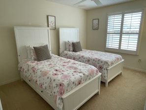 A bedroom with two single beds, white headboards, floral bedding, and a window with blinds.