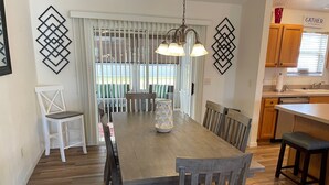 Comedor con mesa de madera, sillas y candelabro. Zona de cocina con armarios de madera y ventana.