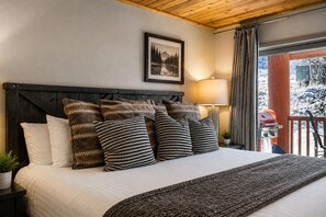 A bed with patterned pillows, a framed picture on the wall, a lamp, and a view of a snowy landscape through a window. - 51 Snowflower 2 Bedroom Condo by RedAwning (Park City)
