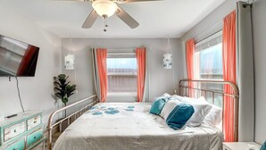 A bedroom with a bed, a ceiling fan, a TV, a dresser, and a window with curtains.