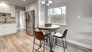 A modern kitchen with white cabinets, a stainless steel refrigerator, a round dining table with four chairs, and a window with blinds.