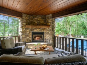 Una chimenea de piedra con fuego, una terraza de madera, mobiliario exterior y vistas a un cuerpo de agua.