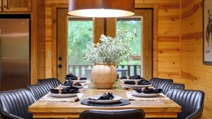 A wooden interior with a dining table set for four, a large vase with greenery, and a hanging light fixture.