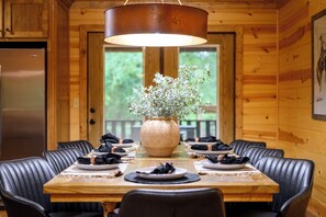A wooden interior with a dining table set for four, a large vase with greenery, and a hanging light fixture.