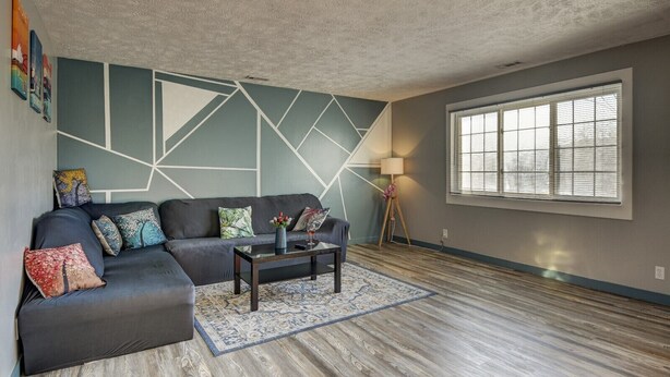 Un salon avec un canapé gris foncé, une table basse et un tapis à motifs. Il y a une fenêtre avec des stores et une lampe sur un trépied.