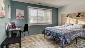 Une chambre à coucher avec un lit, un bureau, une chaise, une fenêtre et un mur orné d’un tableau.