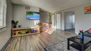 Un vaste espace de vie doté d’un plancher en bois, d’un téléviseur fixé au mur, d’une bibliothèque remplie de livres et d’une plante, ainsi que d’un canapé sur lequel repose un verre de vin sur une table d’appoint.