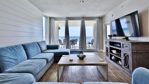 A living room with a sofa, a coffee table, and a TV on a wooden cabinet. There is a balcony with a view of the sea.