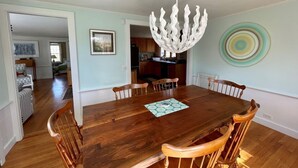 A dining area with a wooden table and chairs, a modern chandelier, and a kitchen in the background. - Cozy Chatham home with privacy and nautical decor (Chatham)
