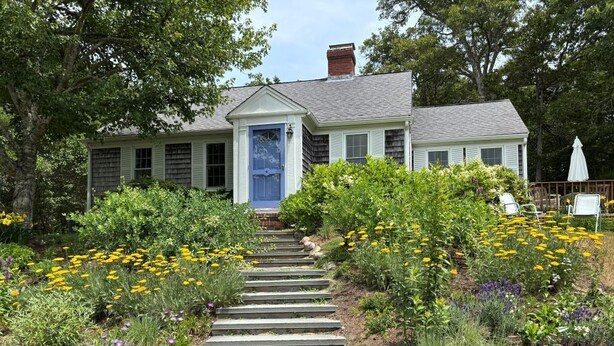 A house with a blue door, surrounded by a garden with yellow and purple flowers, and a stone staircase leading to the front door. - Cozy Chatham home with privacy and nautical decor (Chatham)