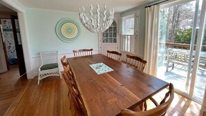 A dining room with a wooden table and chairs, a chandelier, and a view of the outdoors through large windows. - Cozy Chatham home with privacy and nautical decor (Chatham)
