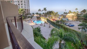 Vista desde un balcón con vista a una piscina, palmeras y un estacionamiento con coches.