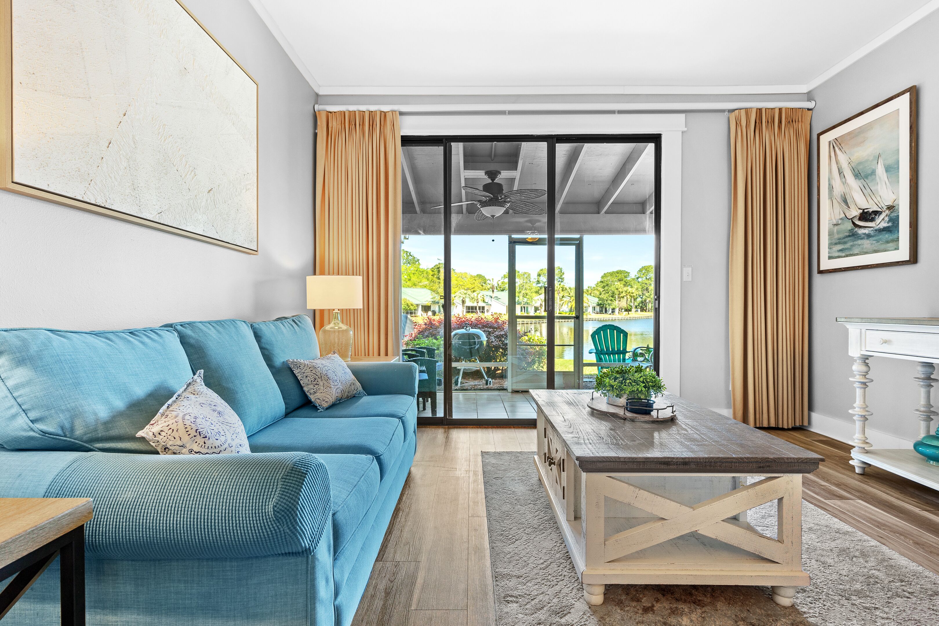Een woonkamer met een blauwe bank, een houten bijzettafeltje en uitzicht door een glazen deur op een buitenruimte met groen.