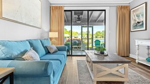 A living room with a blue sofa, a wooden coffee table, and a view through a glass door to an outdoor area with greenery.