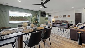 Ein modernes Wohnzimmer mit Essbereich, Deckenventilator und großem Fenster mit Blick auf ein bewaldetes Gebiet.