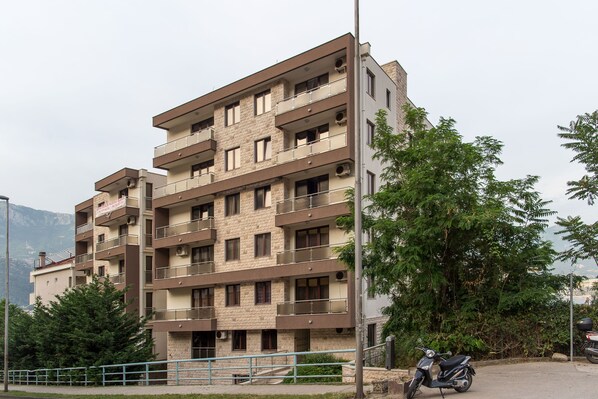 A multi-story residential building with balconies, a paved ground floor, and a scooter parked in front.