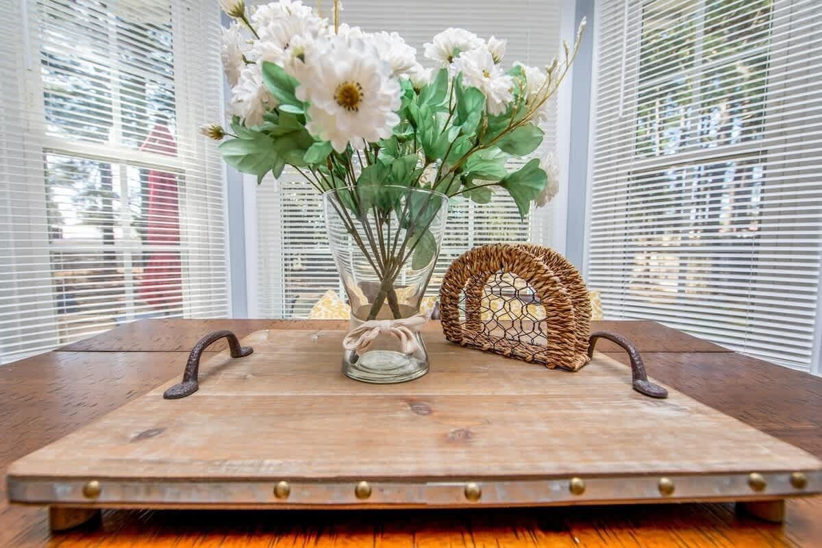 Una mesa de madera rústica con un florero de vidrio con flores blancas, una cesta de mimbre y una bandeja metálica con asas.