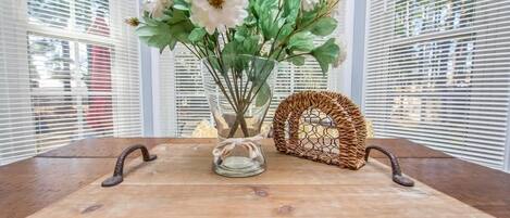 Una mesa de madera rústica con un florero de vidrio con flores blancas, una cesta de mimbre y una bandeja metálica con asas.