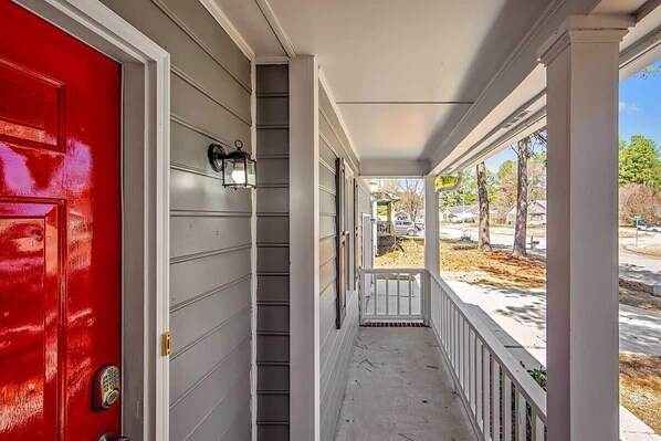 A covered porch with a red door, white railings, and a view of the street.