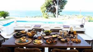 Mesa de café da manhã posta com diversos alimentos e bebidas, com vista para uma piscina e o mar.