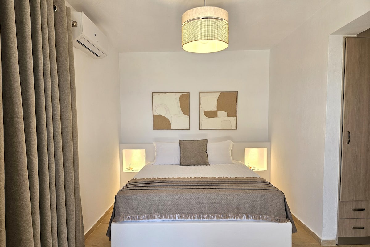 A bedroom with a large bed, two bedside lamps, a ceiling light, and two abstract wall art pieces.