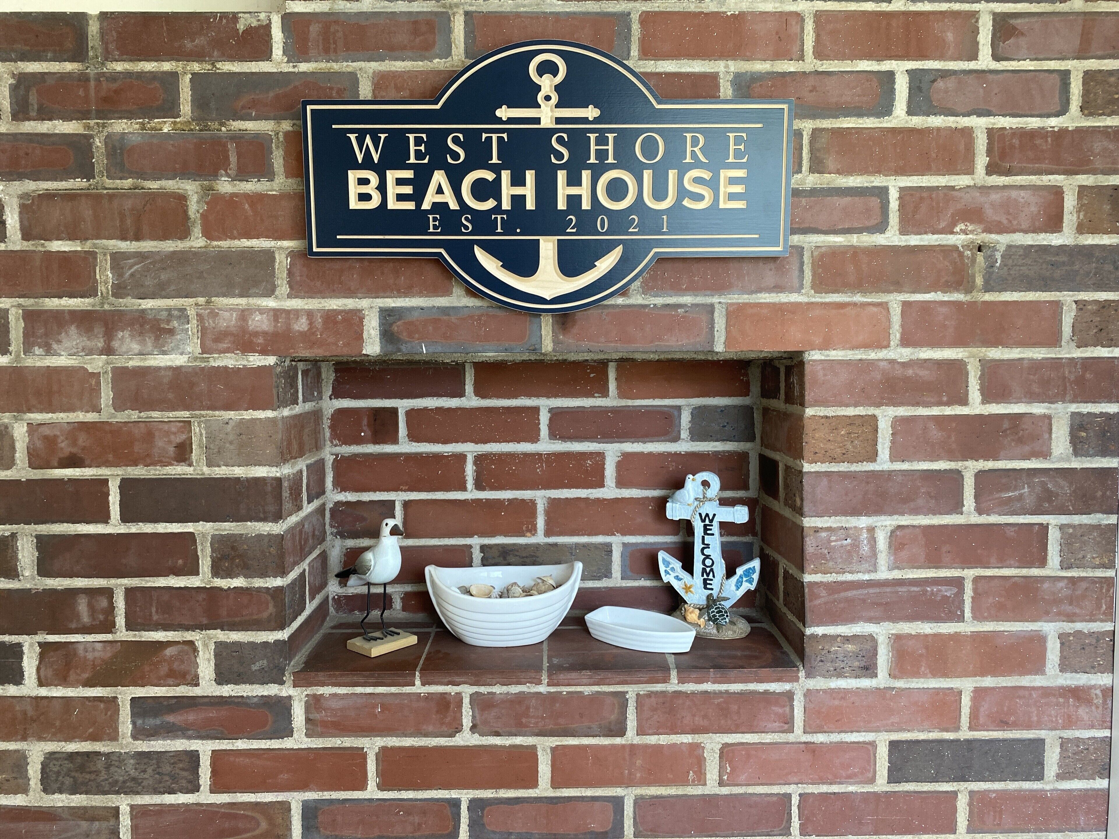 Un muro de ladrillo con un letrero que dice 'West Shore Beach House Est. 2021' y un pequeño estante con adornos.