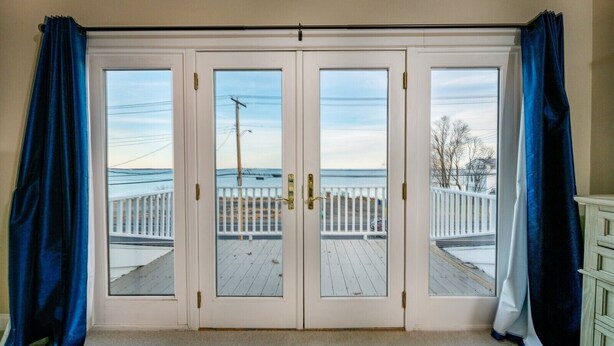 A room with a large sliding glass door leading to a wooden deck with a white railing, overlooking a body of water. - Beautiful, Spacious, Beachfront Home - Perfect for Entertaining Family & Friends (West Haven)