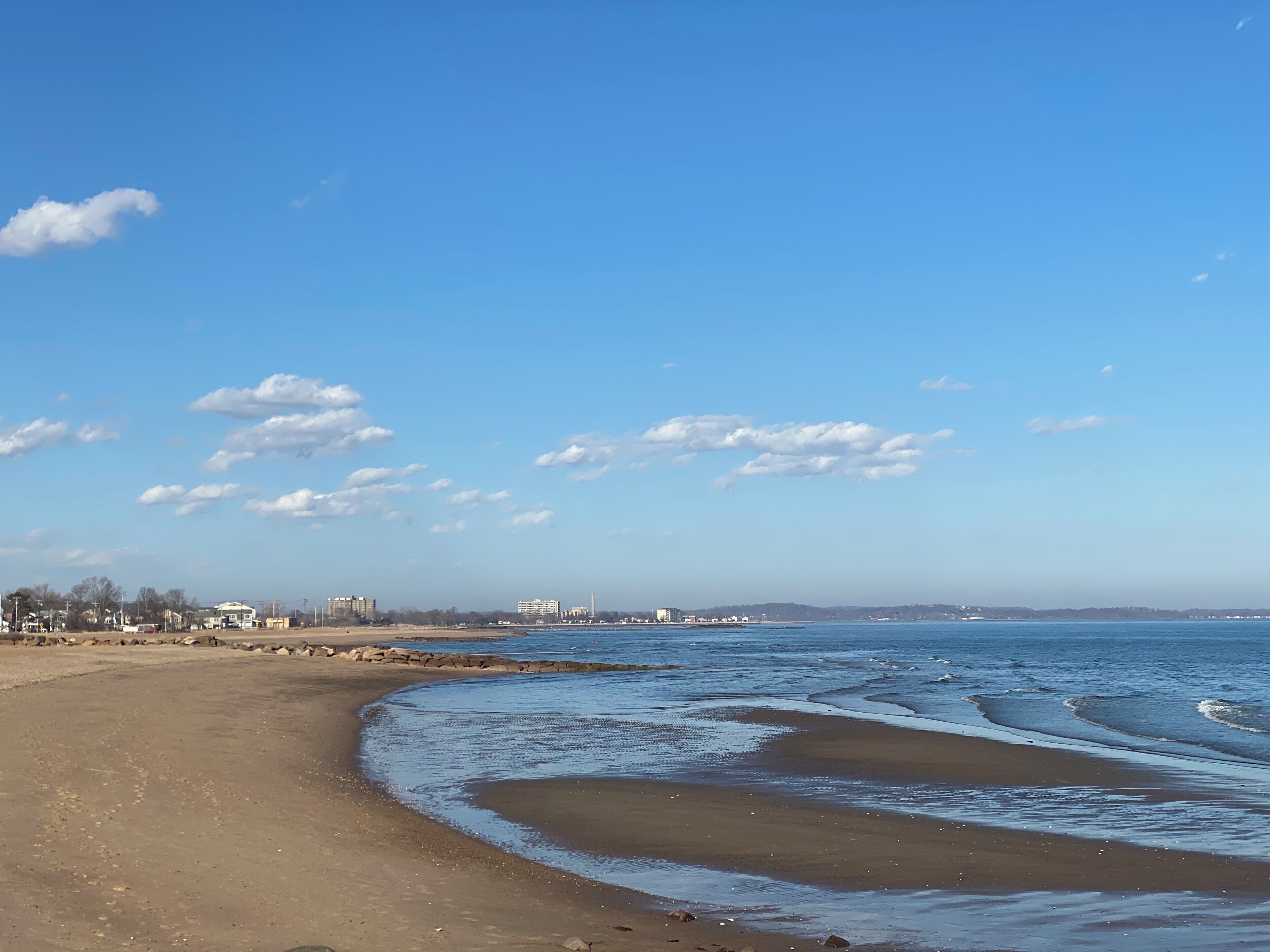 A beach with a clear sky and calm sea. There are buildings in the distance.