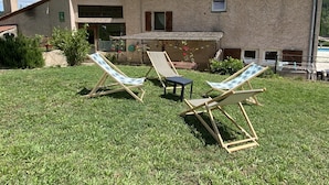 En bygning med et tagdækket tag, en lille patio med bord og stole, og en græsplæne med to liggestole.