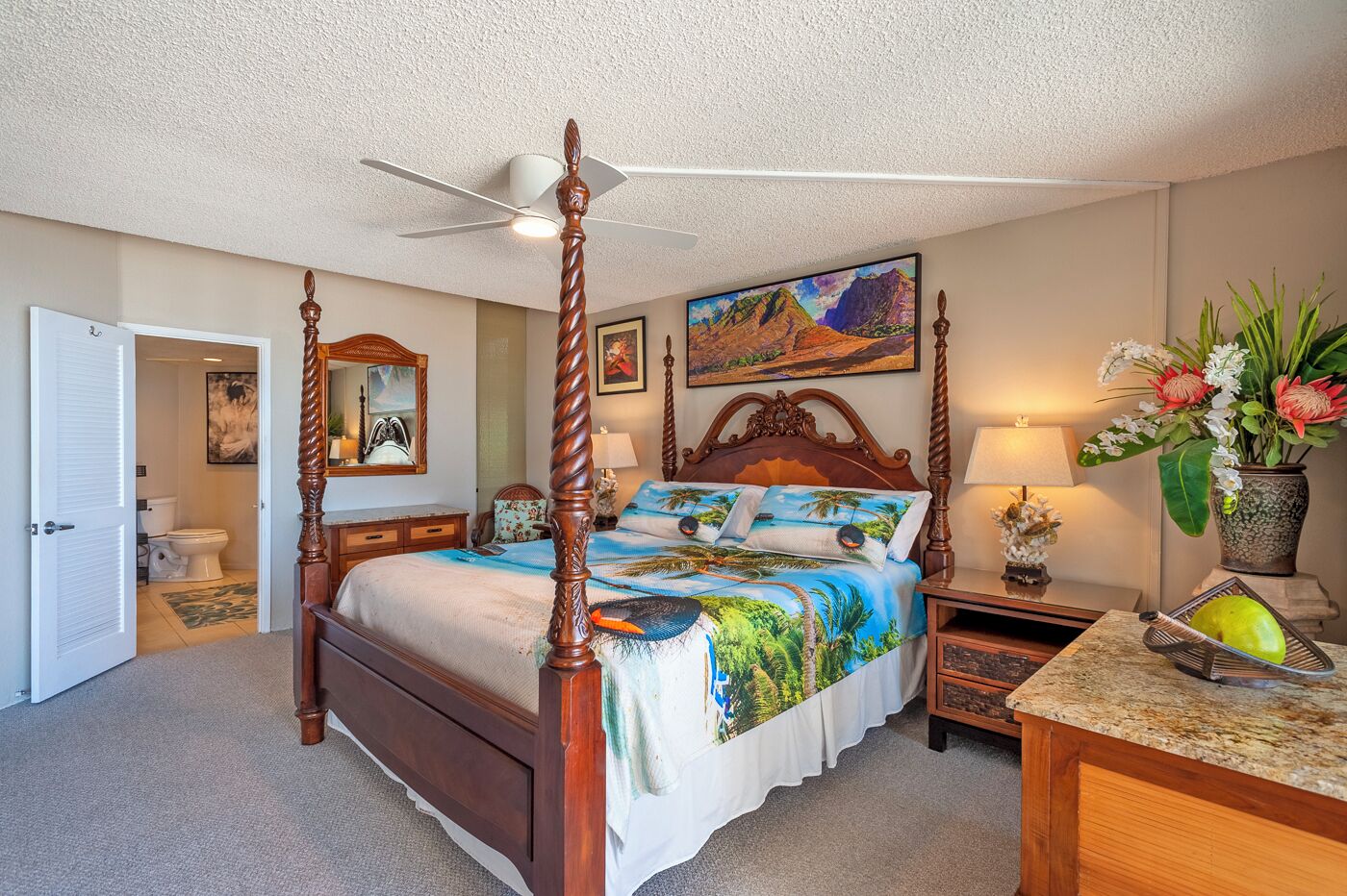 A four-poster bed with a tropical beach design, a ceiling fan, a painting above the bed, and a nightstand with a lamp and a vase of flowers.