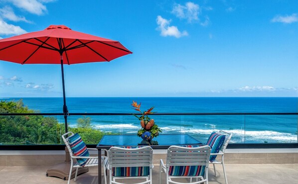 A balcony with a red umbrella, two striped chairs, and a table with a pineapple centerpiece.