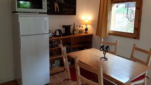 Une cuisine avec un réfrigérateur blanc, un four à micro-ondes, une table en bois avec des chaises, une étagère contenant des ustensiles de cuisine et une fenêtre avec des rideaux.