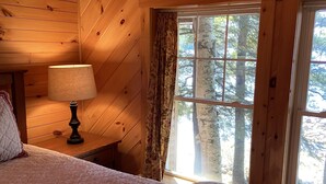 Dormitorio de cabaña de madera con cama, mesita de noche, lámpara y ventana con cortinas.