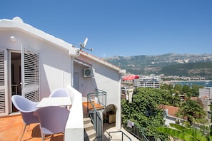 A balcony with a white table and chairs, a red umbrella, and a view of the sea and mountains. - Panoramic apartment (Budva)