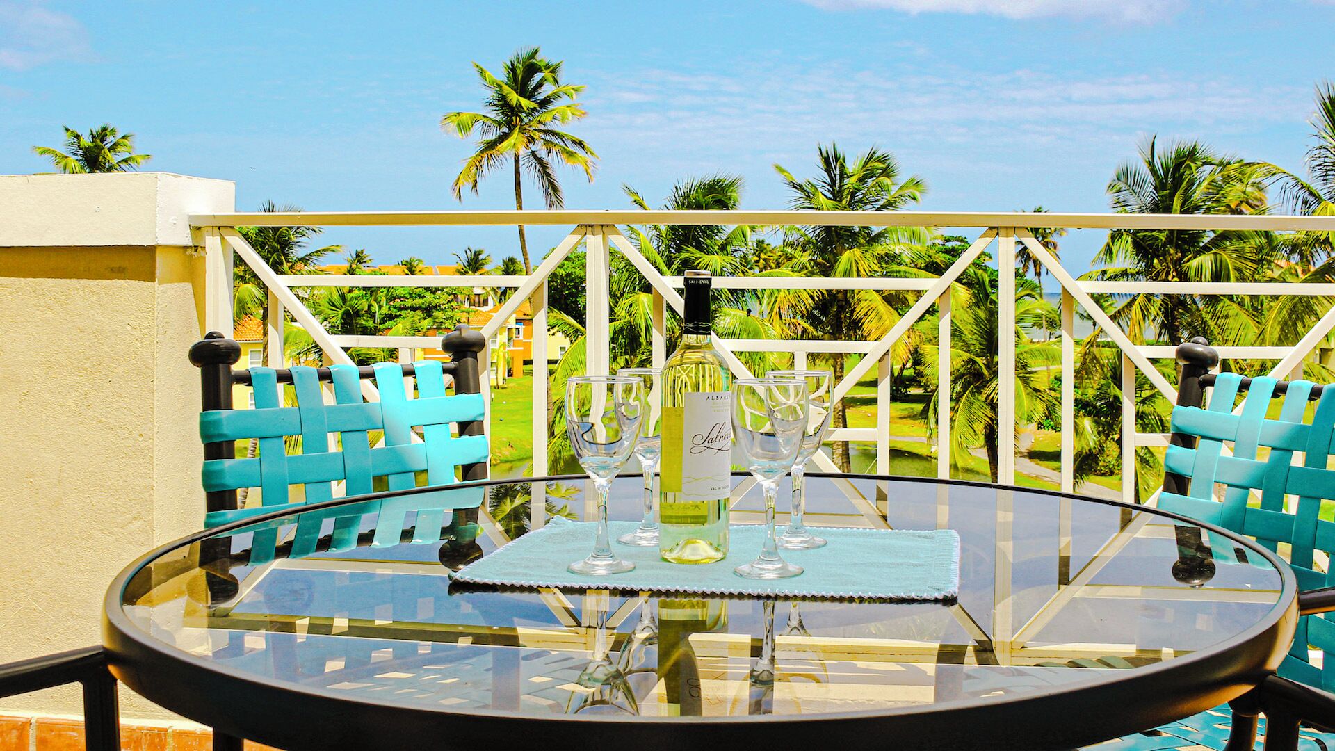 A round glass table with a bottle and glasses, surrounded by blue chairs, set against a backdrop of palm trees and a clear sky.