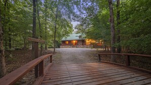 A wooden bridge leading to a rustic cabin with a stone foundation, surrounded by dense forest. - Holiday Road! Cabin with Beautiful Outdoors, Game Room and Hot Tub (Broken Bow)